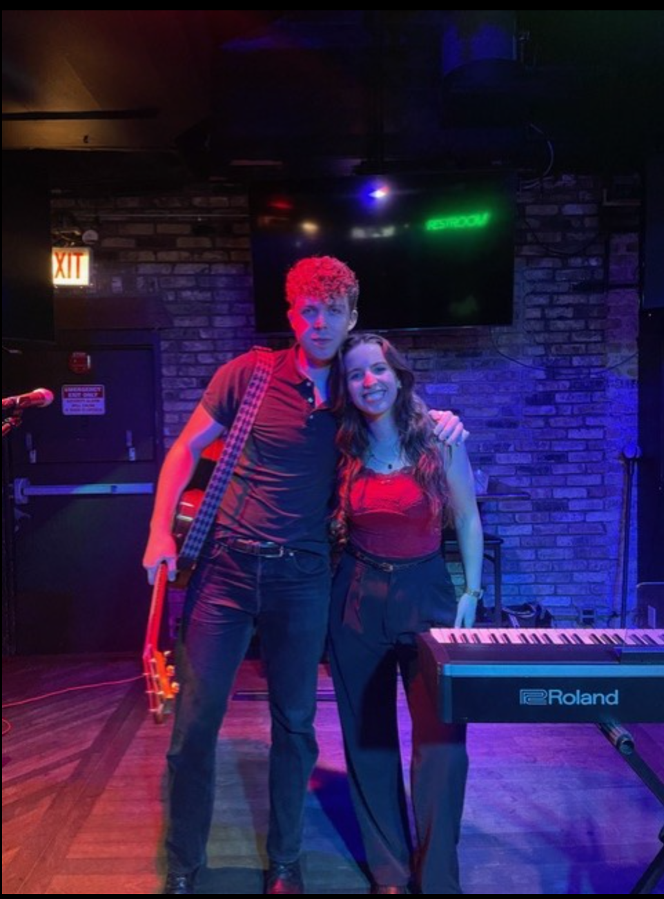 A man holding a guitar taking a picture with a woman near a electric piano under purple light